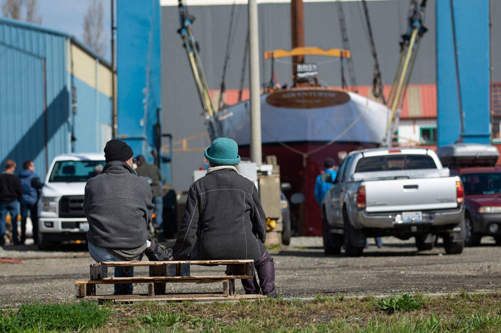 Two people sitting on pallets watch as the schooner Adventuress is lifted Friday. (Jesse Major/Peninsula Daily News)