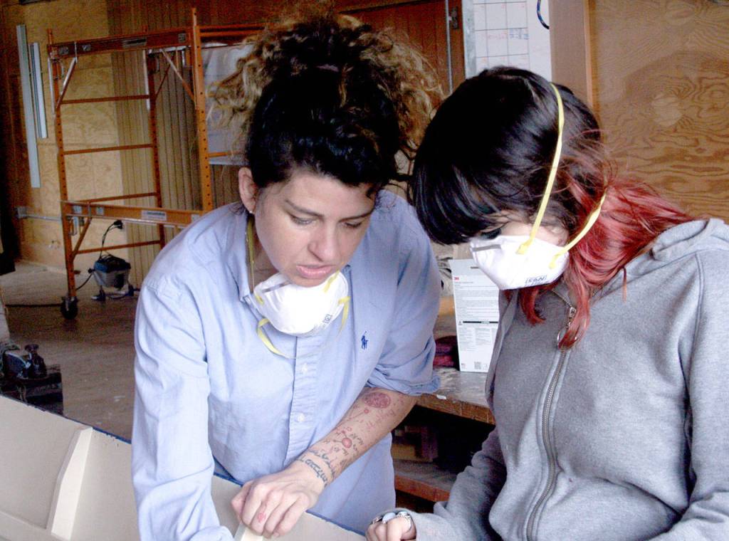 Instructor Ariela Marshall teaches Kira McDowell how to use sandpaper with the grain as they work on a wooden boat Wednesday at the Northwest Maritime Center. (Brian McLean/Peninsula Daily News)