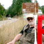 Norm Norton of Eaglemount sits in a chair in front of the Anderson Lake State Park sign along state Highway 20 on Monday. The protest group from the Jefferson County Environmental Coalition added a sign that reads poison above the fish on the state park sign. (Brian McLean/Peninsula Daily News)