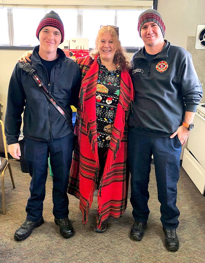 Quilcene Fire Rescue firefighters Nick Singleton, left, and Zak Torres stand with Brinnon and Quilcene Community Knitters founder Lise Solvang while wearing knit caps that the knitters made and donated to first responders. (Brinnon and Quilcene Community Knitters)