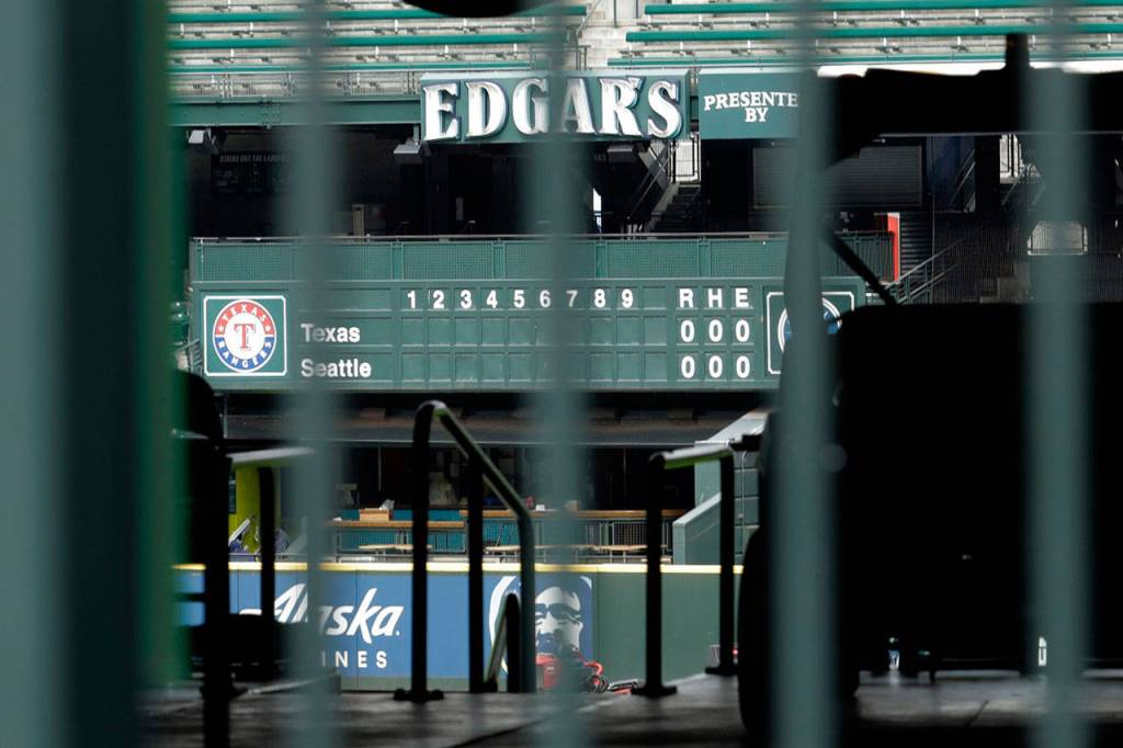 The manual scoreboard at T-Mobile Park in Seattle is seen through the bars of an entry gate Thursday, March 26, 2020, on the day that would have been the Mariners Opening Day baseball game against the visiting Texas Rangers. Earlier in the month, Major League Baseball called off the start of the season due to the outbreak of the new coronavirus. (Ted S. Warren/Associated Press file)