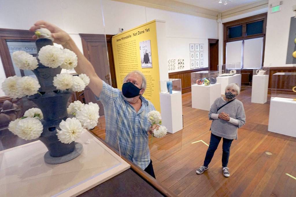 Stephen Yates puts dahlias in a glazed stoneware vase made in 1996 by artist Anne Hirondelle as Hirondelle watches Saturday morning while standing inside her own exhibit at the Jefferson Museum of Art & History on the museums first day open since closing in March. When Hirondelles exhibit first opened March 5 before abruptly closing due to the growing coronavirus pandemic, the vase was filled with tulips. Yates is a member of the museums exhibition committee and helped set up Hirondelles exhibit in March. (Nicholas Johnson/Peninsula Daily News)