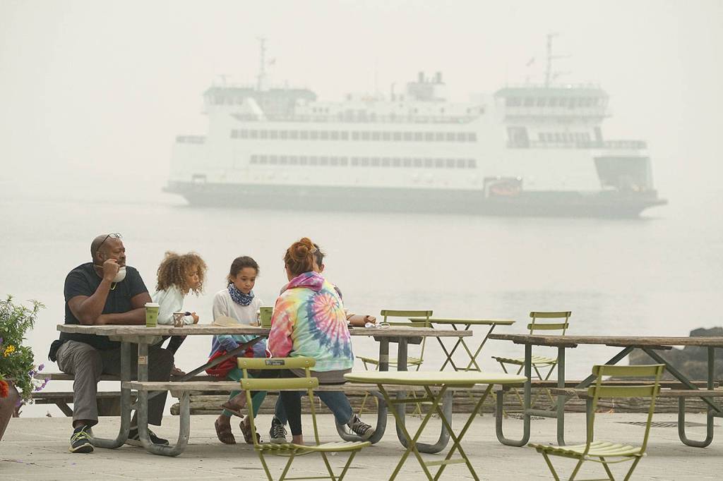 A family visiting Port Townsend from Washougal, which sits on the Washington side of the Columbia River just east of Portland, enjoys coffee and other treats Saturday morning in a downtown pavilion at Water and Tyler streets as a smoke-shrouded MV Kennewick passes behind them in Port Townsend Bay. (Nicholas Johnson/Peninsula Daily News)