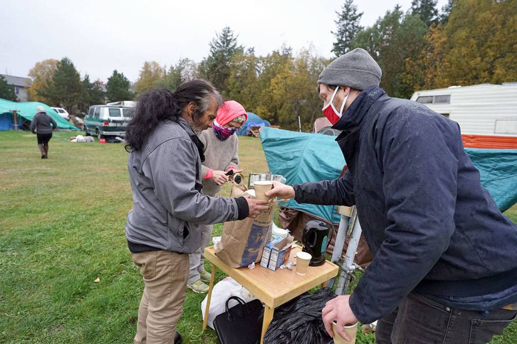 Brian Richardson, program manager at Dove House Advocacy Services Recovery Cafe, right, hands a cup of hot cider to John Hall, left, as Jessica Treibel, center, takes some socks on offer to those living at the Jefferson County Fairgrounds campground as rain falls Tuesday morning in Port Townsend. (Nicholas Johnson/Peninsula Daily News)