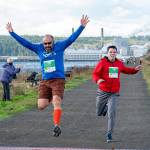 Sequim’s Russ Britton (507) jumps through the finish line after he completes the Run the Peninsula 5K on the Larry Scott Trail in Port Townsend on Saturday. Taran Johnson, (547) also from Sequim, races to the finish. (Steve Mullensky/for Peninsula Daily News)