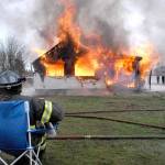 Firefighter/paramedic Erik Sundin sits in a folding chair to watch a house at 2354 E. Fifth Ave., in the Gales Addition east of Port Angeles, go up in flames on Sunday as part of a training exercise for personnel of Clallam County Fire District 2. The donated house was torched to allow firefighters to hone their skills over a two-day session that culminated in destruction of the structure. Once extinguished, the charred remains will be cleared to make way for future development. (Keith Thorpe/Peninsula Daily News)