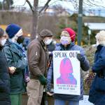 Debbi Steele of Port Townsend shows her sign to Mado Most, far left, and Annalee McConnell just before the candlelight vigil at Pope Marine Park in downtown Port Townsend on Thursday afternoon. About 100 people joined the League of Women Voters-Jefferson County vigil, marking the first anniversary of the insurrection at the U.S. Capitol. Earlier in the day, a vigil in Sequim drew at least 125 people. (Diane Urbani de la Paz/Peninsula Daily News)