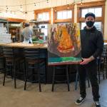 Larry White, district manager for Barhop Brewing & Artisan Pizza and artist, holds a painting for Barhop's new Sequim location.Behind him, kitchen manager Jeffrey Adams prepares pizzas for customers in the open kitchen. (Emily Matthiessen/Olympic Peninsula News Group)