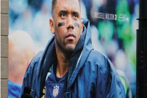 A pedestrian walks past a photo of Seattle Seahawks quarterback Russell Wilson at Lumen Field, Tuesday, March 8, 2022, in Seattle. The Denver Broncos agreed to send a massive haul of players and draft picks to the Seattle Seahawks in a trade for Wilson, two people familiar with the negotiations confirmed to The Associated Press on Tuesday. The blockbuster trade, which is pending Wilson passing a physical, can't become official until the start of the new league year on March 16, 2022. (AP Photo/Ted S. Warren)