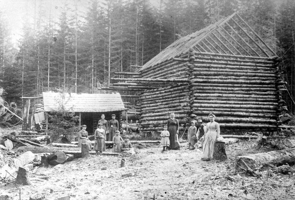 A two story log cabin being built. One man, four women and five children (Unidentified) are shown. A smaller log cabin is beside the cabin under construction. (North Olympic History Center)