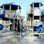 Port Angels Parks & Recreation Department employee Easton Goslin hoses down the concrete underlayment at the playground at Shane Park on the west side of Port Angeles on Wednesday. The playground was closed in mid-September after tiles of rubberized material that made up the play surface began to come loose, creating a hazard to play equipment users. The old tiles have been removed and are awaiting replacement. The playground area remains closed until work can be completed. (Keith Thorpe/Peninsula Daily News)