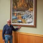 Emily Matthiessen / Olympic Peninsula News Group
Photographer Ross Hamilton stands with one of the 35 prints of his 4-by-5-inch negatives that are displayed in the hallways of the Lodge at Sherwood Village. Hamilton says that each of his photographs has a story behind it; this one was a brief moment in time when the light overwhelmed him with its beauty coming through the trees on the Dosewallips River. The joy of seeing something really beautiful is what makes photography so special, he says.