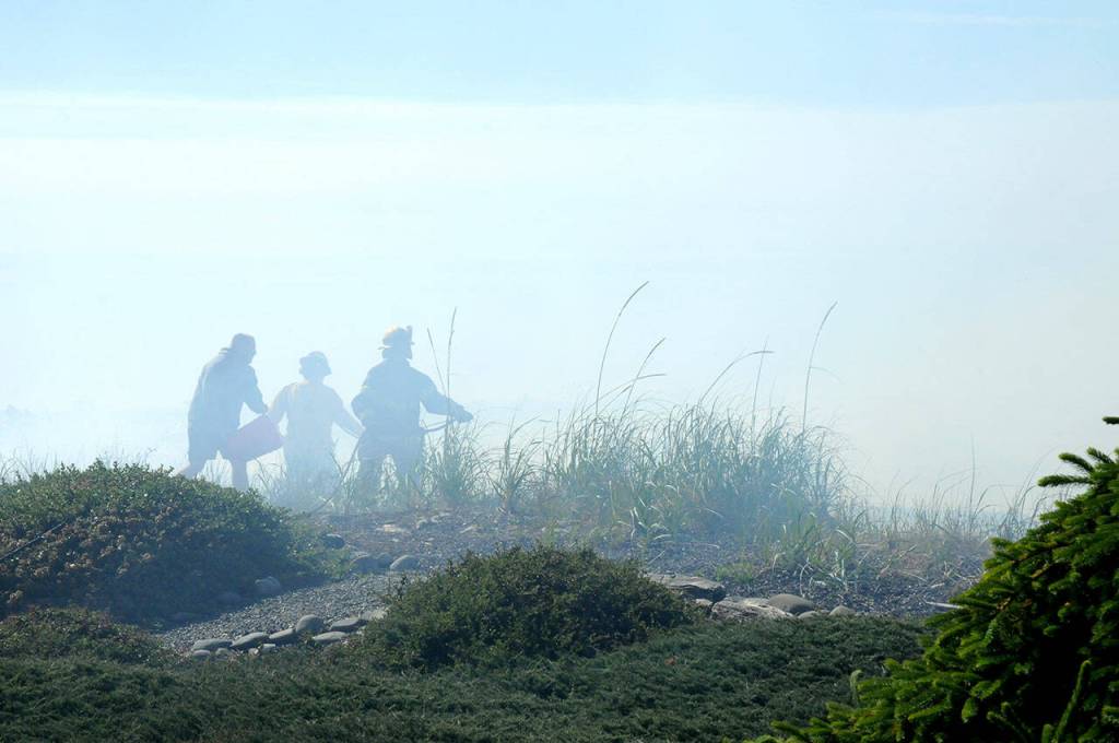 Firefighters receive assistance with a bucket of water from a Four Seasons Ranch resident while fighting a beach fire that threatened homes along the Strait of Juan de Fuca on Saturday. (Keith Thorpe/Peninsula Daily News)