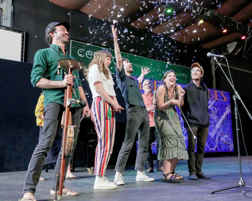 Joel Savoy, left, outgoing Centrum Fiddle Tunes Artistic Director, reacts as a confetti noisemaker goes off behind him after he introduces the group The Onlies as the new artistic director and the first time a group has been selected for that position. The Onlies are a group of old friends who grew up playing fiddles, guitars and banjos in their hometowns of Seattle and Lexington, Va. Next to Savoy are Sami Braman, Riley Calcagno, Vivian Leva and Leo Shannon. Centrum staffer Peter McCracken is behind them. (Steve Mullensky/for Peninsula Daily News)