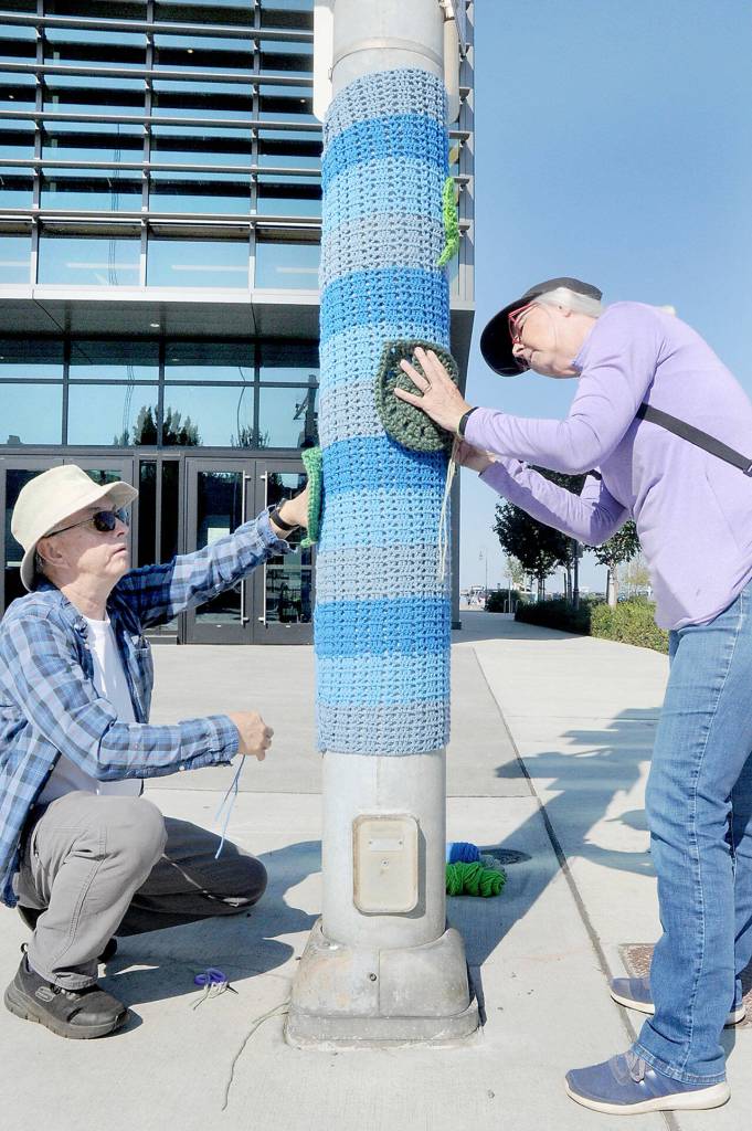 Juan and Joyce Ruiz of Joyce wrap a woven yarn blanket as crocheted decorations around a light pole at First and Oak streets in downtown Port Angeles on Saturday as part of a yarn bombing project commissioned by First Federal in celebration of the financial institutions centennial. Mary Sue, co-owner of Cabled Fiber & Yarn, which produced the wrappings, said they had planned to wrap numerous poles and trees in the 100 block of West Front Street and surrounding streets and that the yarn project would last as long as they last. (Keith Thorpe/Peninsula Daily News)