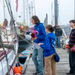 Ocean Smith gets ready to hand off a vase of flowers to a Northwest Maritime Center sailing team member already aboard during an early Friday morning delivery of flowers purchased on Thursday. Looking on are teammates Ava Butterfield and Charley Capel. (Steve Mullensky/for Peninsula Daily News)