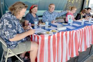 Pie contest judges, from left, Jeff Tocher, Laurel Hargis and Clallam County Commissioner Randy Johnson, along with event sponsor Country Aire Natural Foods representatives Kristina Fallon, Katie Meyers and Caeron Alarcon, make their choices during Thursdays annual Independence Day apple pie contest at Port Angeles City Pier. A total of 26 pies were entered into the contest with first-place honors going to Xaven McCarty of Seattle, Elisabeth Pennell of Mukilteo in second and Selena Reach of Port Angeles taking third. The contest was dedicated the late legacy judge and former Peninsula Daily News publisher John Brewer, represented by a photo in front of an empty chair. (Keith Thorpe/Peninsula Daily News)