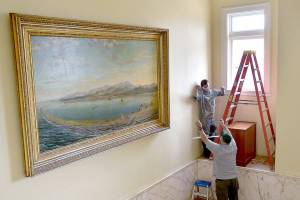 David Brownell, executive director of the North Olympic History Center, top, takes a piece of ultraviolet-filtering window tinting from Ralph Parsons, Clallam County maintenance worker, in an effort on Tuesday to protect historic paintings on the stairway of the section of the county courthouse, including an 1890s depiction of Port Angeles Harbor by artist John Gustaf Kalling. The history center is working with the county to preserve the stairway artworks by adding the window coatings to reduce damage from sunlight and installing an electronic UV monitor to track potentially harmful rays. (Keith Thorpe/Peninsula Daily News)