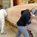 From left to right, Northwest School of Wooden Boatbuilding students Krystol Pasecznyk and Scott McNair sand a Prothero Sloop with Sean Koomen, the schools boat building program director. Koomen said the sanding would take one person a few days. He said the plan is to have 12 people sand it together, which will take a few hours. (Elijah Sussman/Peninsula Daily News)