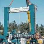 Still suspended and held in steady by a heavy lift crane, a 50-ton crossmember beam of the new 300-ton variable width Travelift acquired by the Port of Port Townsend is bolted down by an employee, lower right, on Monday. (Steve Mullensky/for Peninsula Daily News)