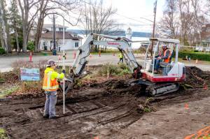 Workers from Van Ness Construction in Port Hadlock, one holding a grade rod with a laser pointer, left, and another driving the backhoe, scrape dirt for a new sidewalk of civic improvements at Walker and Washington streets in Port Townsend on Thursday. The sidewalks will be poured in early February and extend down the hill on Washington Street and along Walker Street next to the pickle ball courts. (Steve Mullensky/for Peninsula Daily News)