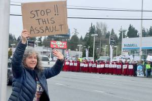 Wendy Rae Johnson waves to cars on the north side of U.S. Highway 101 in Port Angeles on Saturday during a demonstration against U.S. Immigration and Customs Enforcement operations in Minnesota. On the other side of the highway is the Peninsula Handmaids in red robes and hoods. (Dave Logan/for Peninsula Daily News)