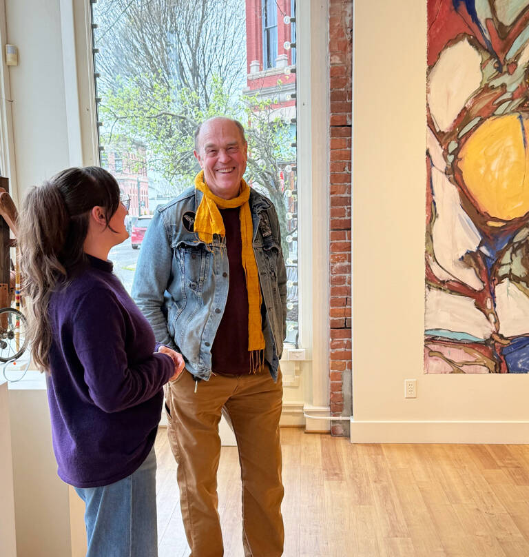 At Northwind Arts Jeanette Best Gallery in Port Townsend, staff member Abby Abramczyk talks with Randy Sturgis, who will do a Q&A about his artwork in the gallery on Saturday. His large-scale paintings include Lundquist Fall Division, partially visible here. (Diane Urbani/Northwind Art)