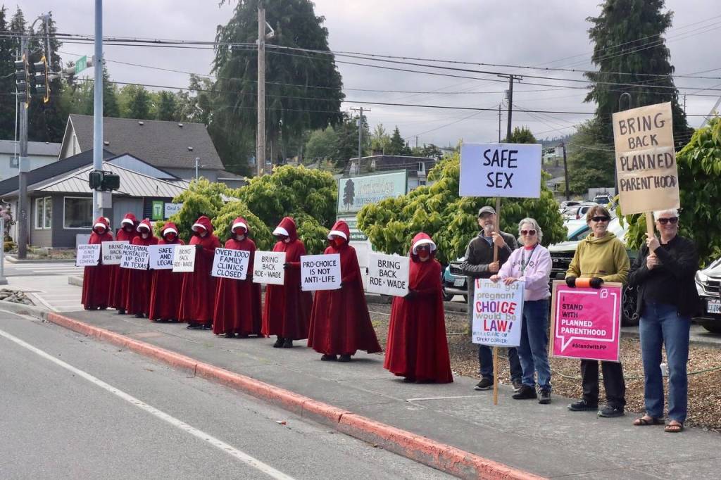 Demonstrators, some dressed as Handmaids, gather at the corner of Eighth and Race streets in Port Angeles on Wednesday. The group holds signs in support of Planned Parenthood, whose clinic on Eighth Street closed in April, and pro-choice messages. (Dave Logan/for Peninsula Daily News)